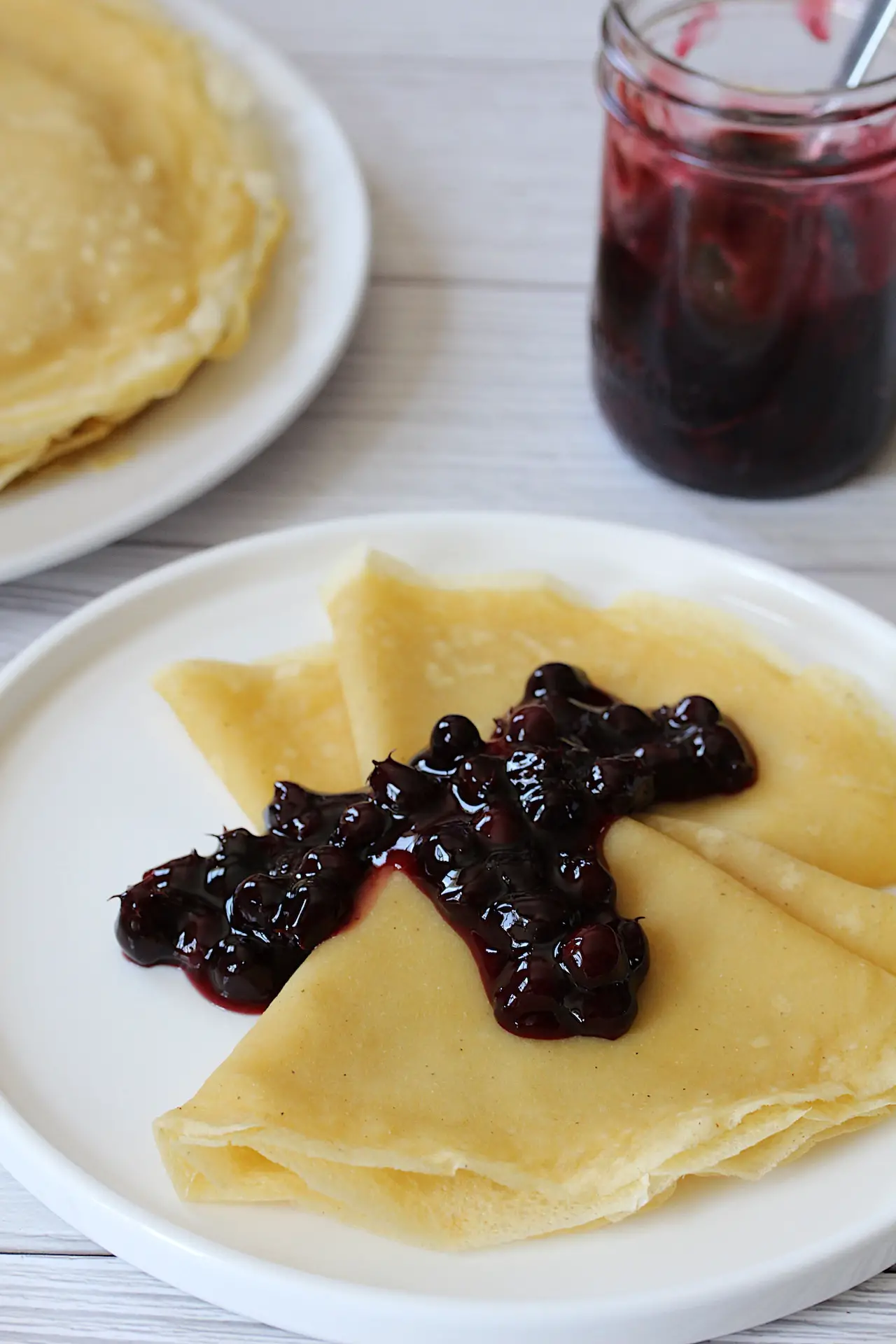 Image of folded crepes on a white plate with Saskatoon berry sauce. In the background is the jar of Saskatoon berry sauce and the plate of stacked crepes.